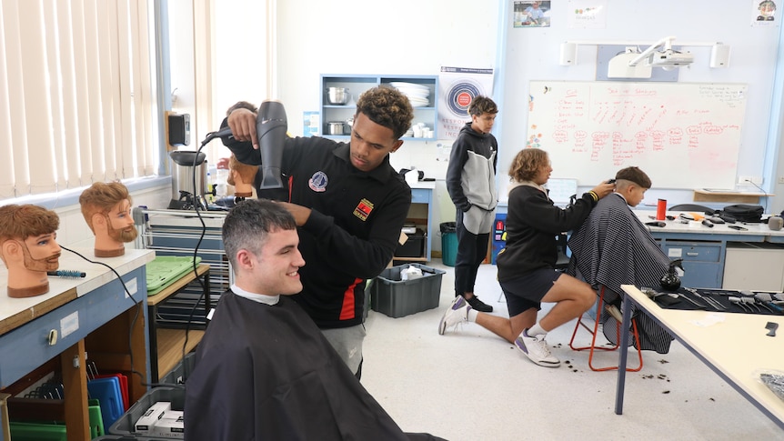 Students blow-drying his teachers hair. Teachers sat in char with black apron around him.