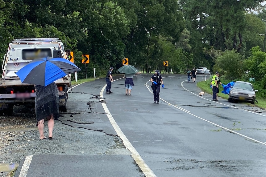 A small brown sedan sits on the side of the road, as emergency workers and other people with umbrellas look around the scene.
