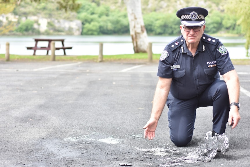 Police officer kneels on the ground in a car park where soot from a fire is evident.