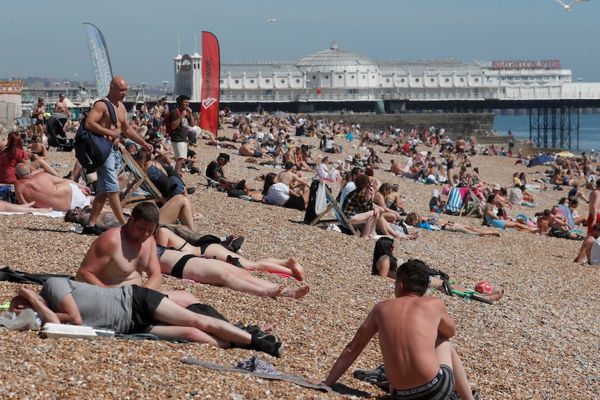 A crowd of people sit on beach on sunny day with water and a pier behind them.