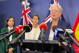 A woman wearing glasses speaks to the media at a press conference as two other women watch in the background.