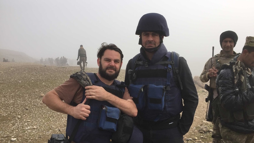 Brown in helmet and safety vest standing next to Hollett holding camera with soldiers in the background.