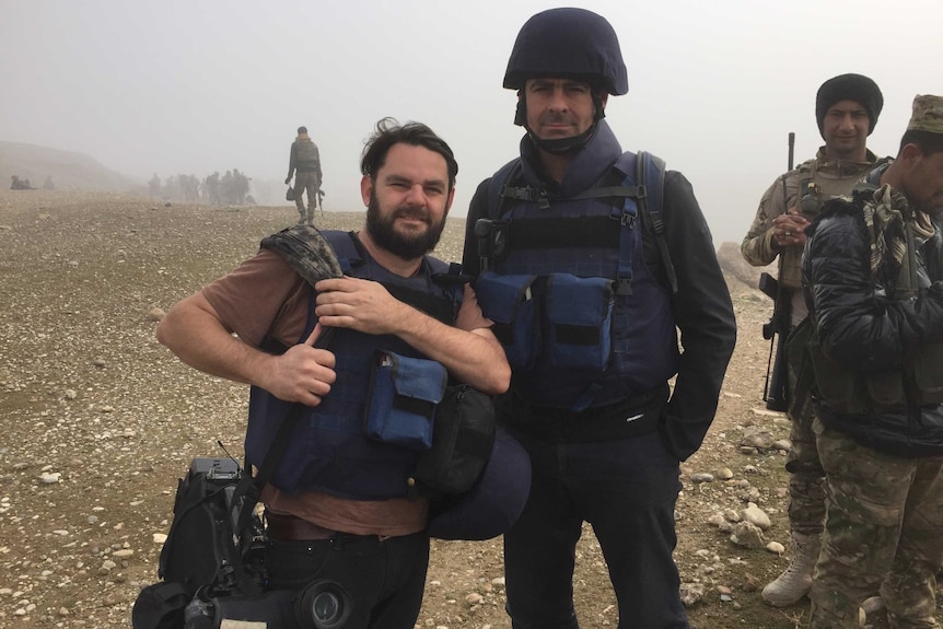 Brown in helmet and safety vest standing next to Hollett holding camera with soldiers in the background.