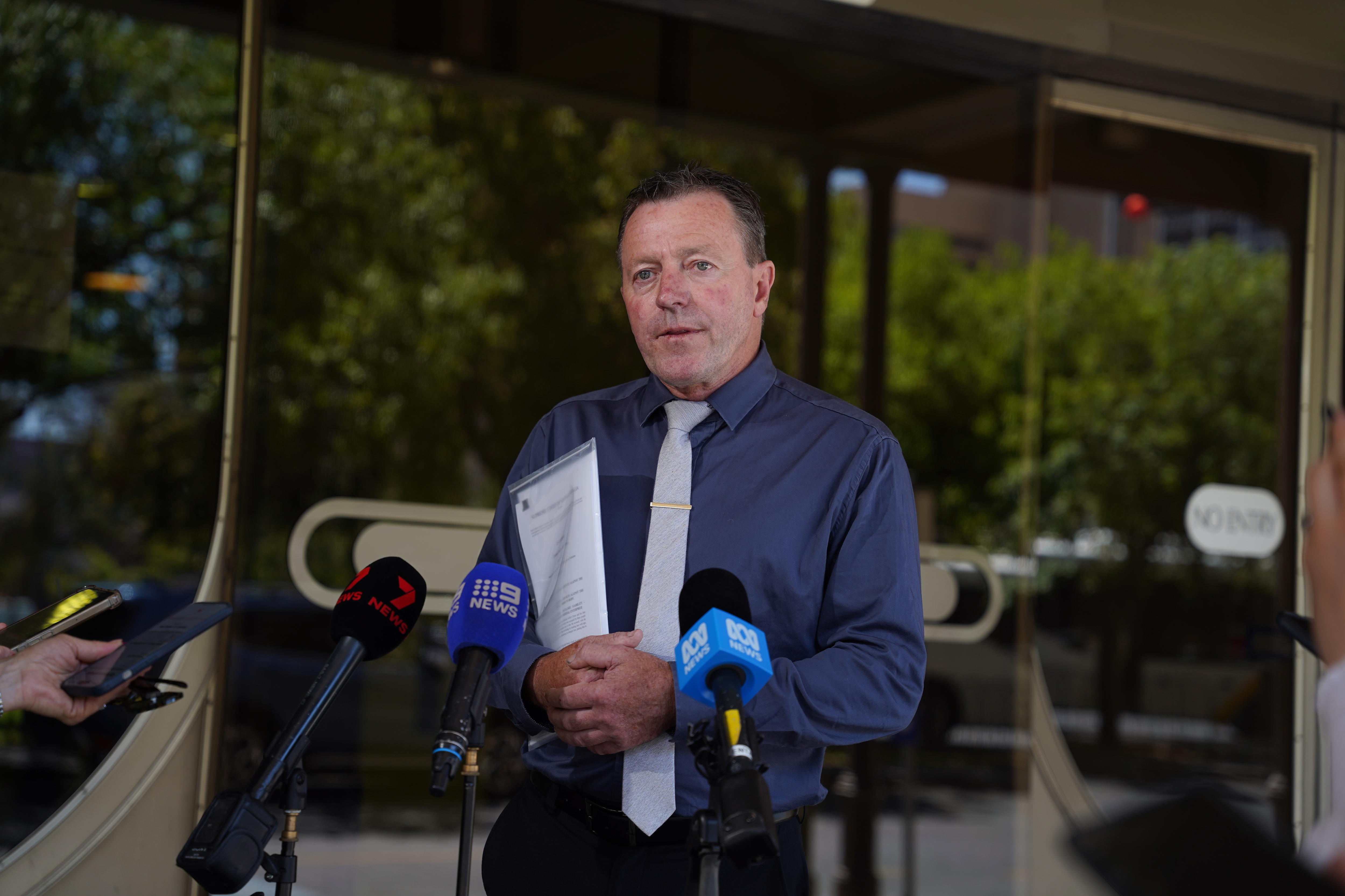 Un hombre de mediana edad, de cabello oscuro, con camisa y corbata, se encuentra frente a un edificio y habla con los medios.