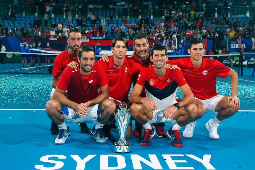 A group of Serbian tennis players in red shirts crouch next to a silver trophy