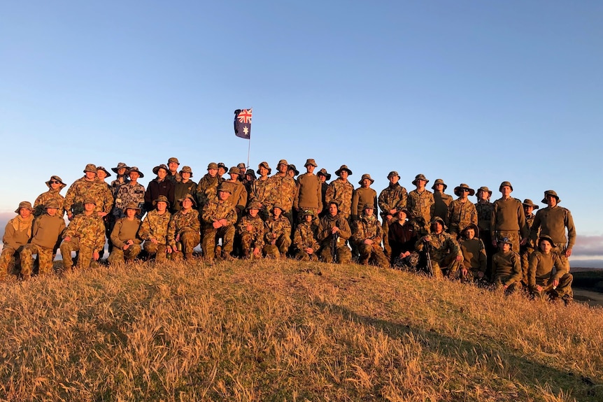 A group of army cadets in uniform standing on a hill with an Australian flag behind them.