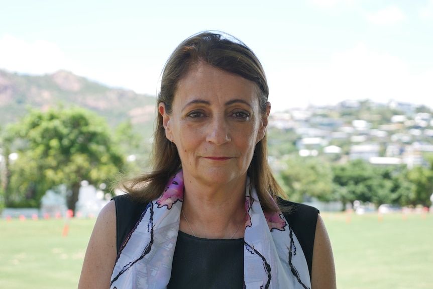 A middle aged woman looks sternly at the camera while stood on a sports field in front of hills.
