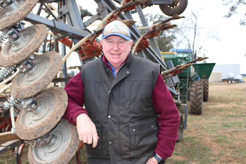 An older man in warm clothes standing in front of farm machinery.