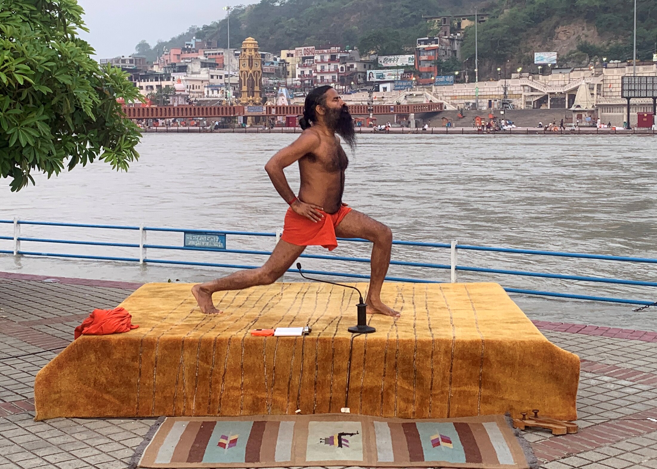 A shirtless Indian man with a long beard does yoga on a platform beside a large river.