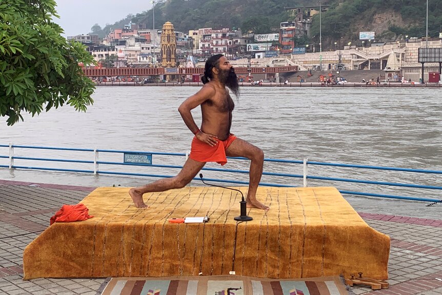 A shirtless Indian man with a long beard does yoga on a platform beside a large river.