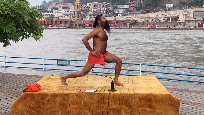 A shirtless Indian man with a long beard does yoga on a platform beside a large river.