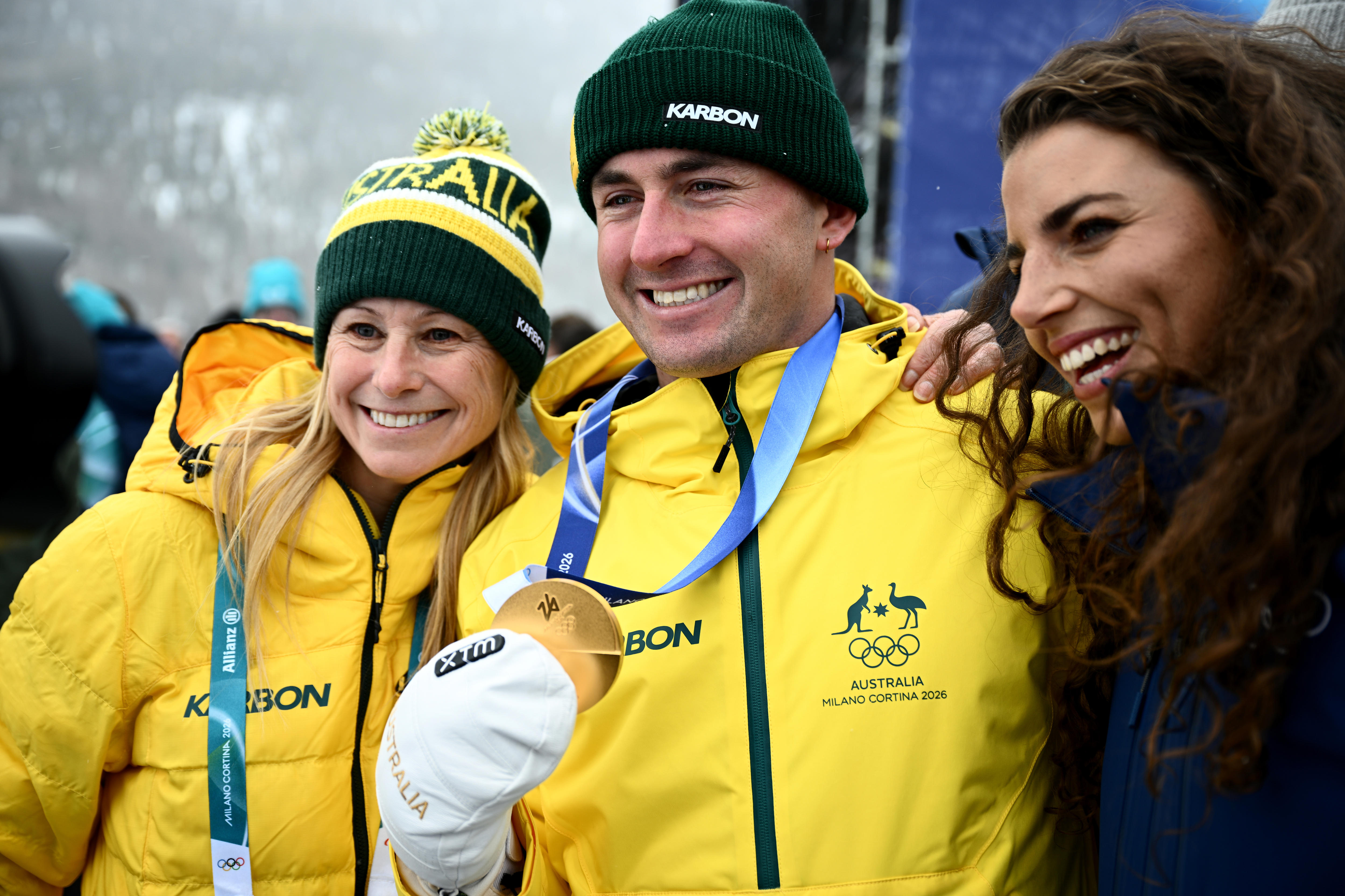 Dos mujeres sonrientes y un hombre sosteniendo una medalla de oro sonríen en la nieve.