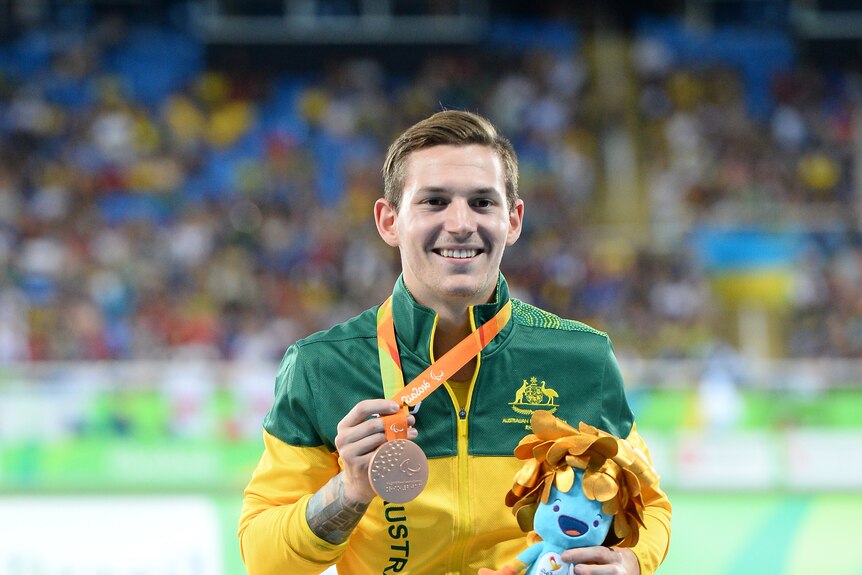 A smiling young man in an Australian tracksuit holds up his Bronze medal and carries the Rio mascot character in other hand