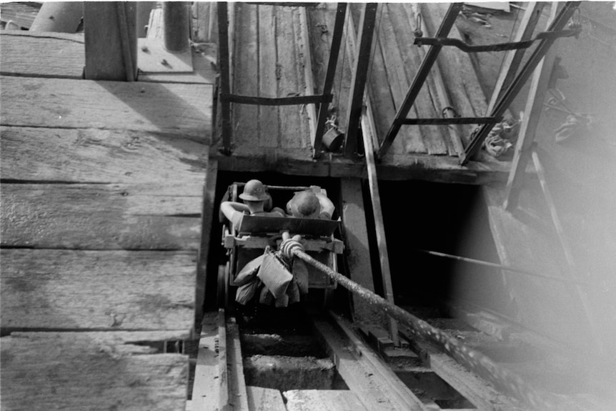 An old black and white photograph of two miners wearing helmets and descending into the shaft in a cart 