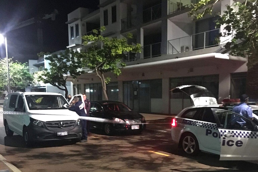 A police car and a white van stand parked on a road outside a three-storey block of units cordoned off by police tape.