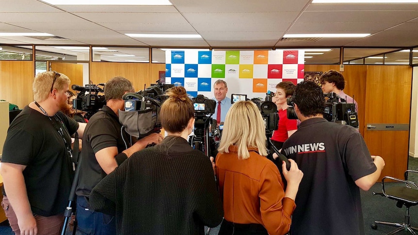A group of journalists and cameramen stand in a semi-circle interviewing the Bundaberg Mayor in his office