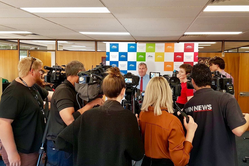 A group of journalists and cameramen stand in a semi-circle interviewing the Bundaberg Mayor in his office