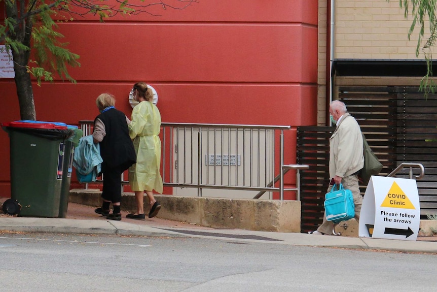 An older woman is escorted into Joondalup Private Hospital by a medical worker in a plastic gown as a man in a facemask follows.