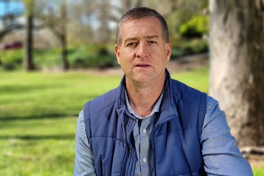 A man wears a blue vest over a blue shirt in a park.