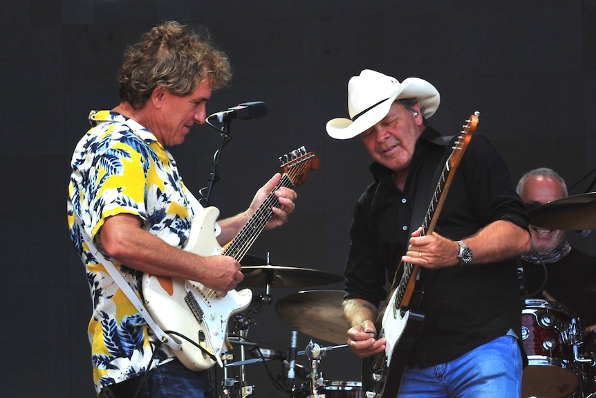 Two men play guitars facing each other on a stage