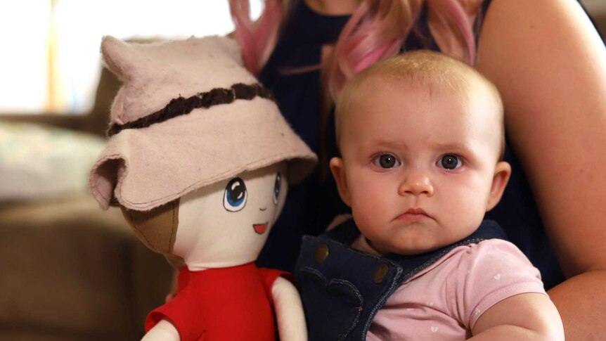 A baby girl holds doll while on mum's lap.