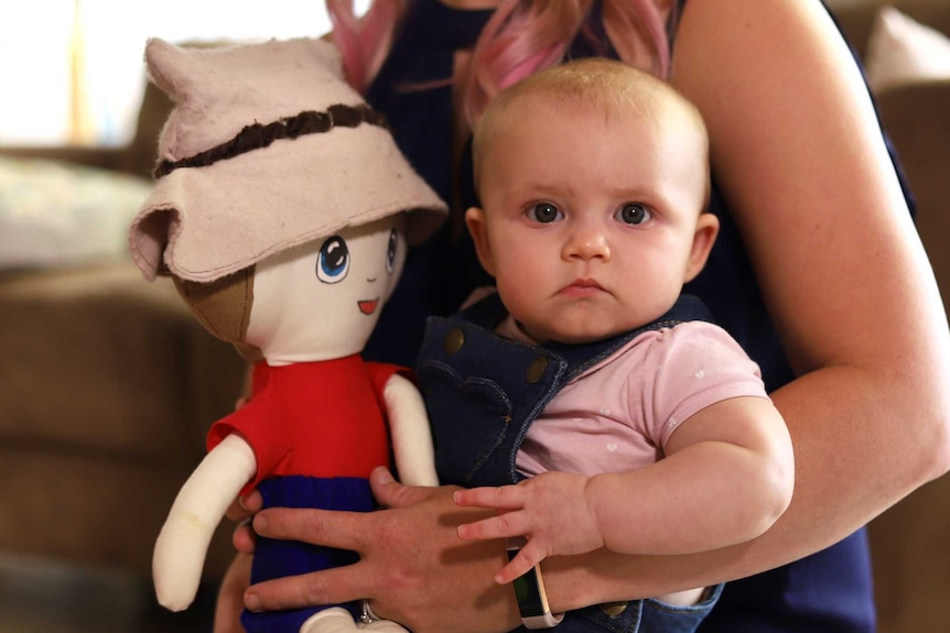 A baby girl holds doll while on mum's lap.