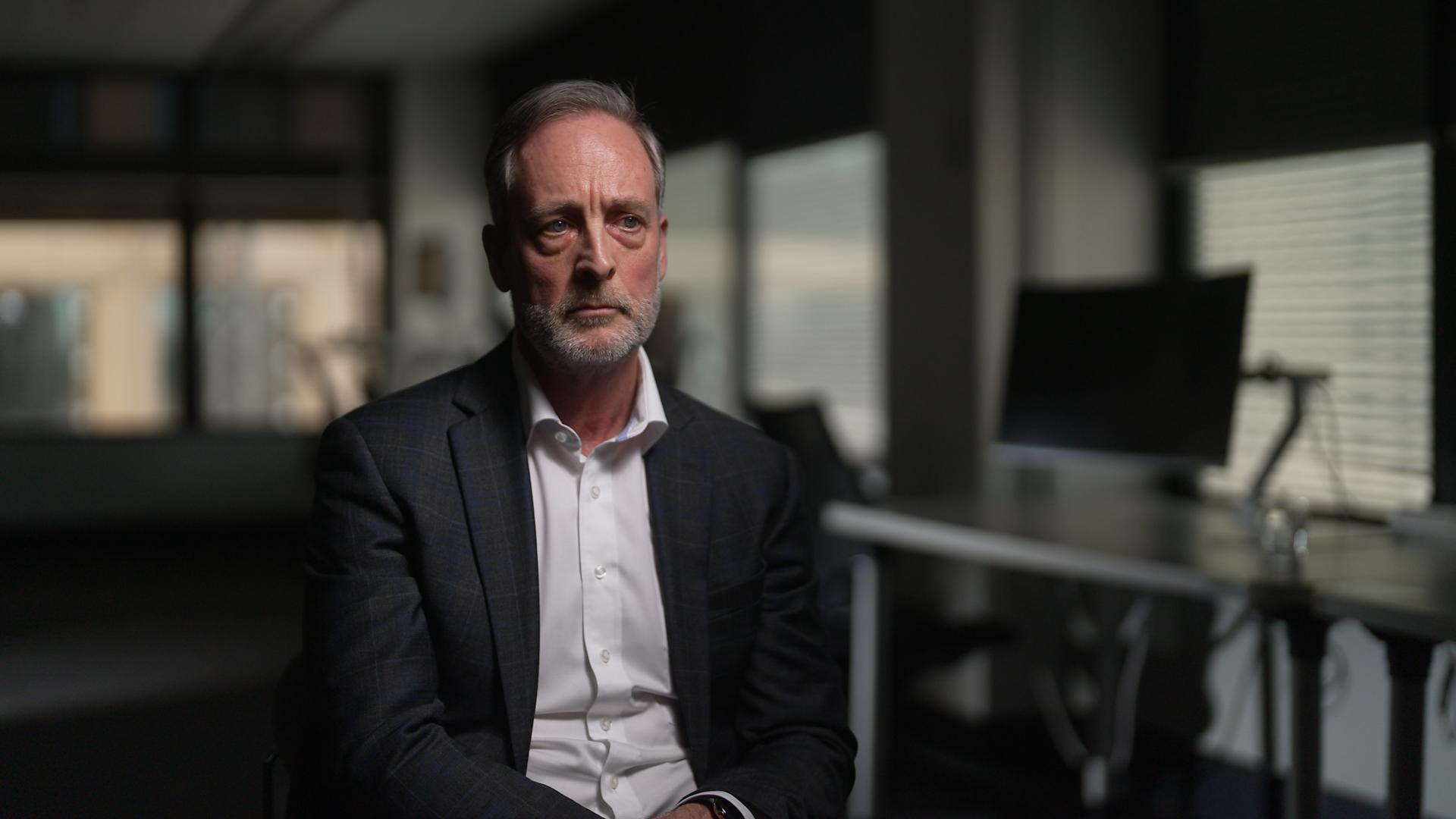 A man in a business shirt and suit jacket sits in a chair in an office, looking serious as he's interviewed for television.