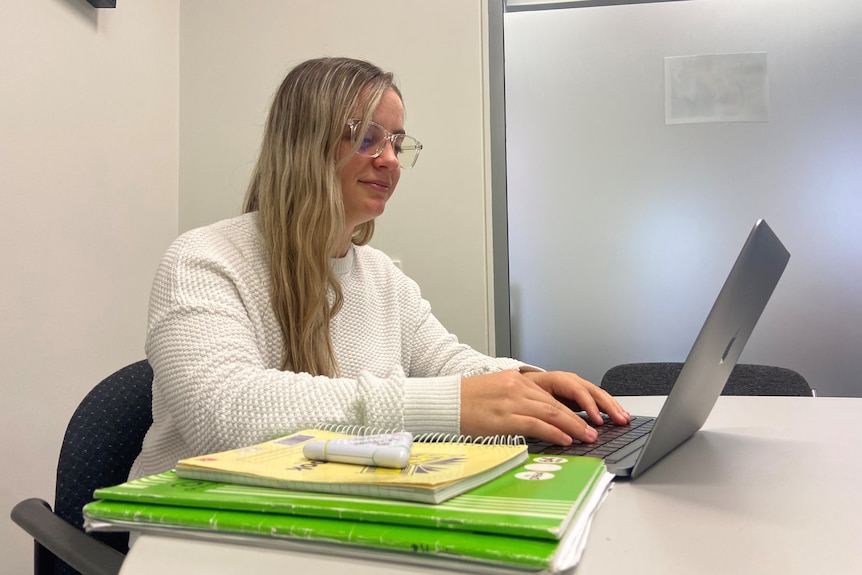 A young woman with long, blonde hair, wearing a white jumper, works on a laptop, with books beside her.