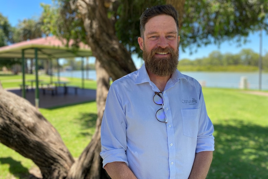 A man in a light blue shirt standing in front of a tree.