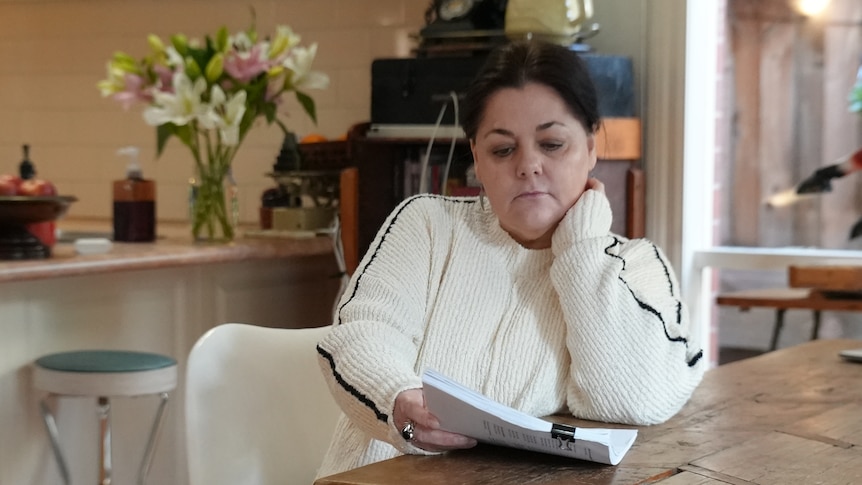 Judy wearing a white sweater, reading a document at her kitchen table.
