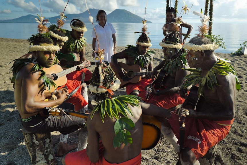 Group of men sitting in a circle, playing guitars and ukeles at the beach and wearing traditional Pacific islander costume. 