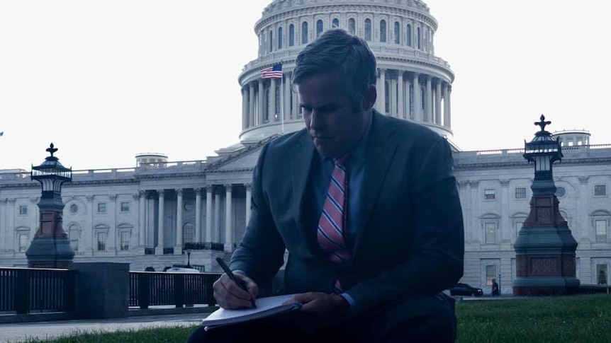 Duffy sitting on camera case on lawn outside Capitol Building writing on notepad.