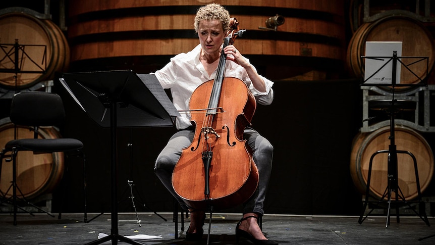 Taryn Fiebig performs cello in front of a giant wooden wine barrel with smaller wine barrels around her.