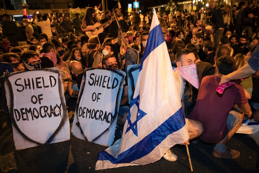 Protesters carrying sings reading "shield of democracy" in English sit near another holding an Israeli flag