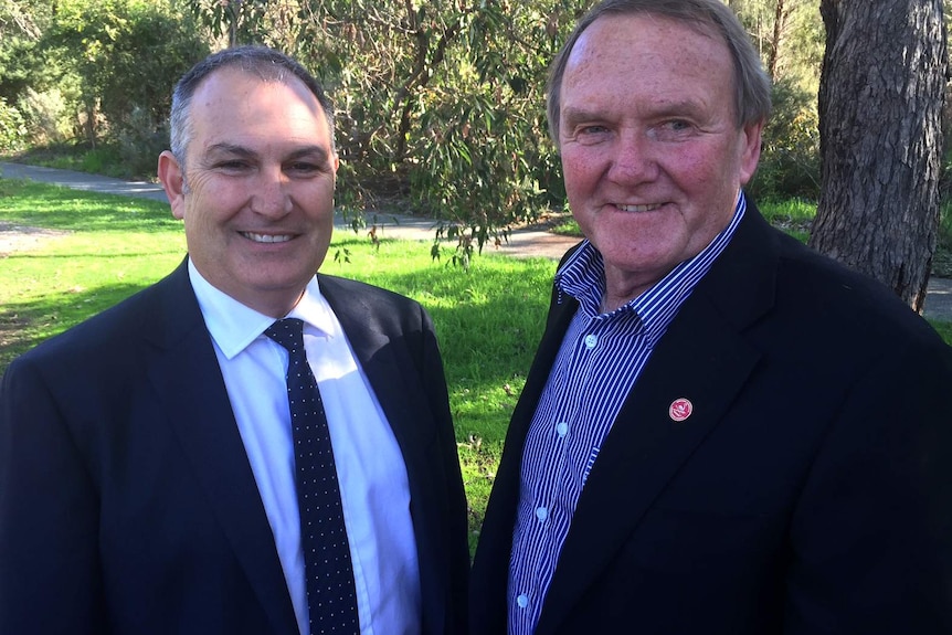 Rick Mazza and Nigel Hallett pose for a photo smiling and wearing suits in front of grass and tree.