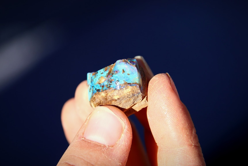 Fingers hold a sparkling blue and green rock opal. 