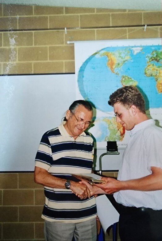 Two men, one older and one younger, with a world map on the wall in the background.