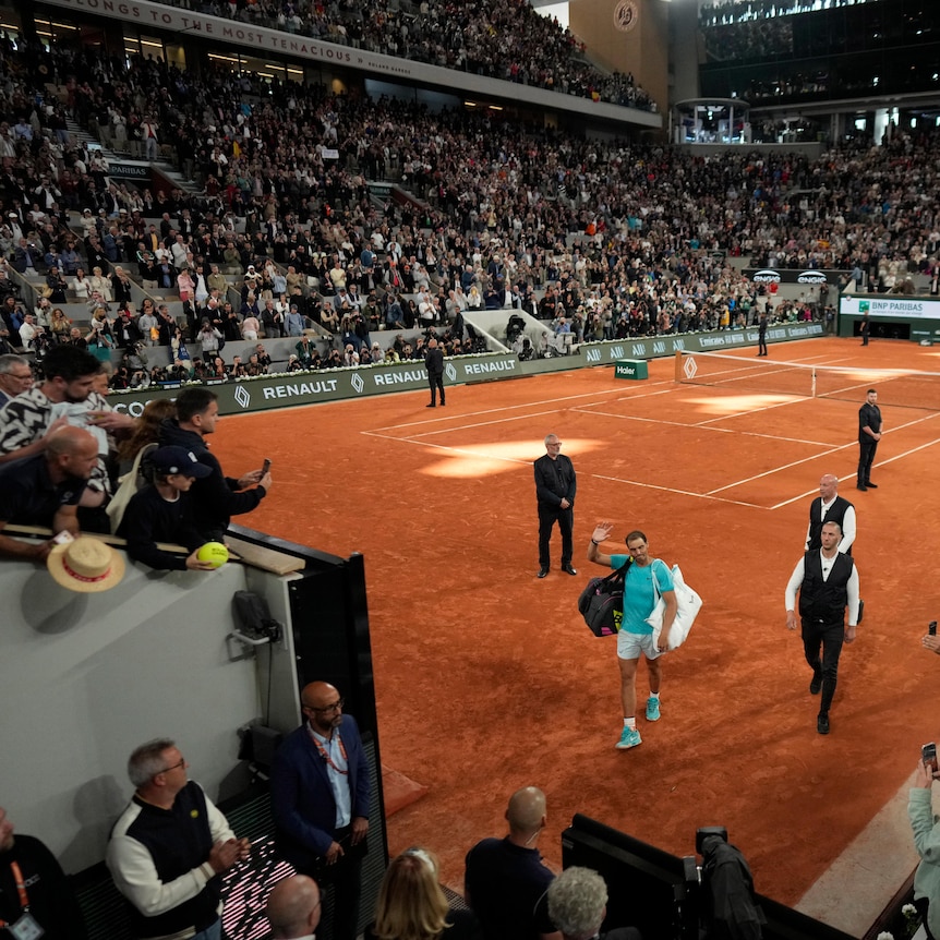 A wide shot of Rafael Nadal waving to the French Open crowd as fans applaud him as he leaves the Roland Garros court.