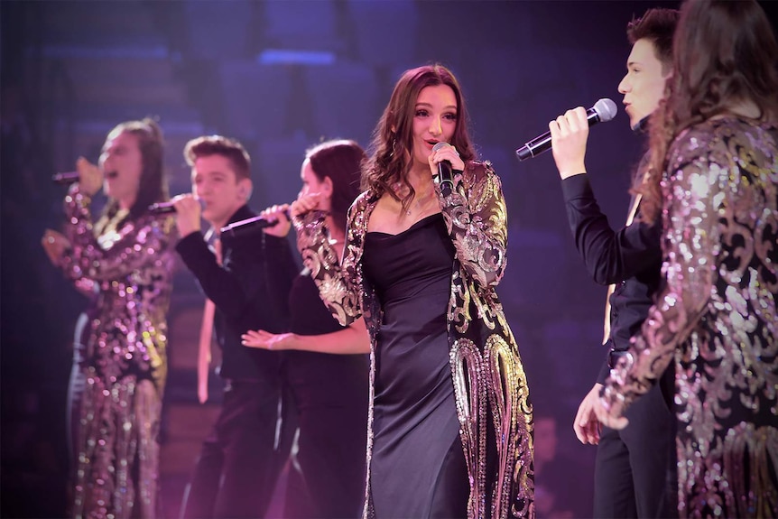 Ella Perusco sings into a microphone alongside other singers on a stage.