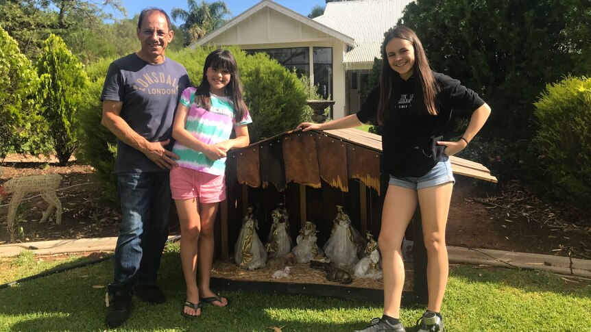 A man and his two daughters pose next to a handmade nativity scene.