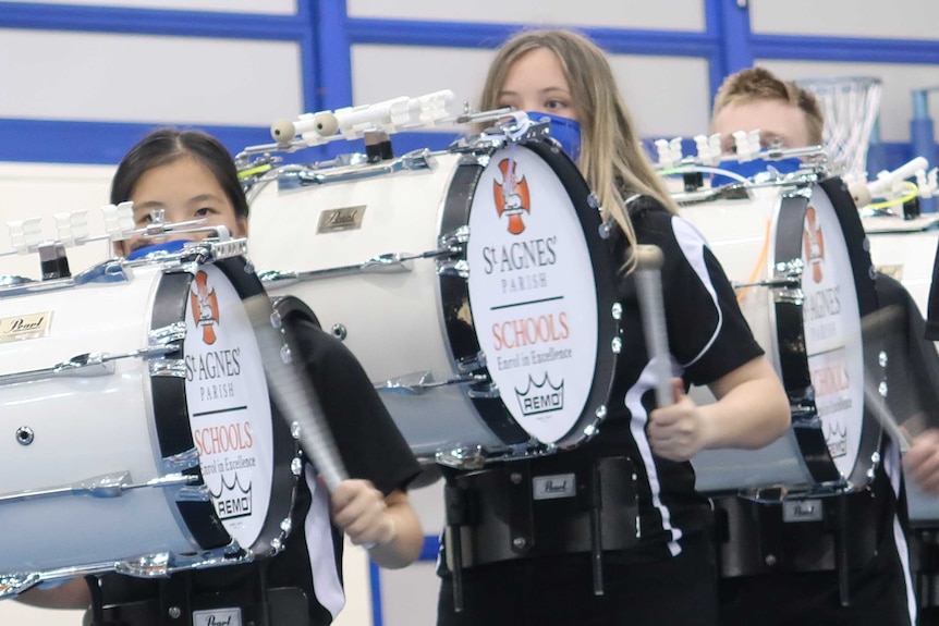 Three students in black t-shirts stand in line with their eyes barely seeing over bass drums strapped to their bodies.