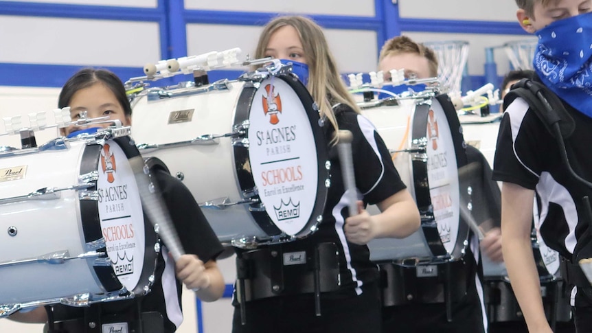 Three students in black t-shirts stand in line with their eyes barely seeing over bass drums strapped to their bodies.
