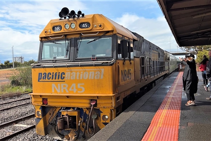 A diesel train with the words Pacific National on the front arrives at a station. A woman in black beam and waves at it.