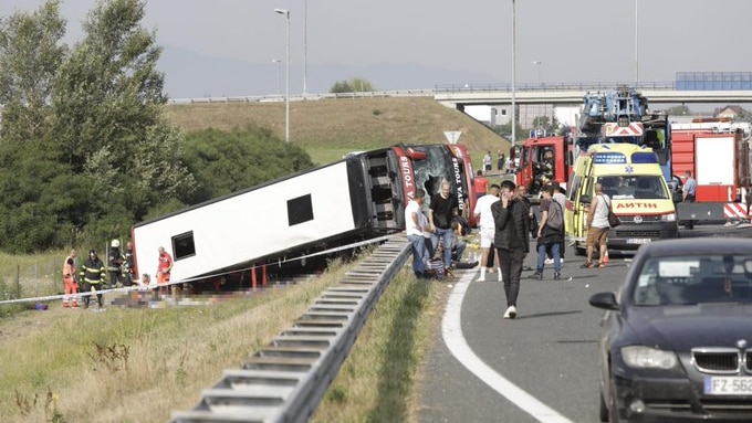 A white bus is toppled over on its side next to highway as rescue workers surround it on a sunny day