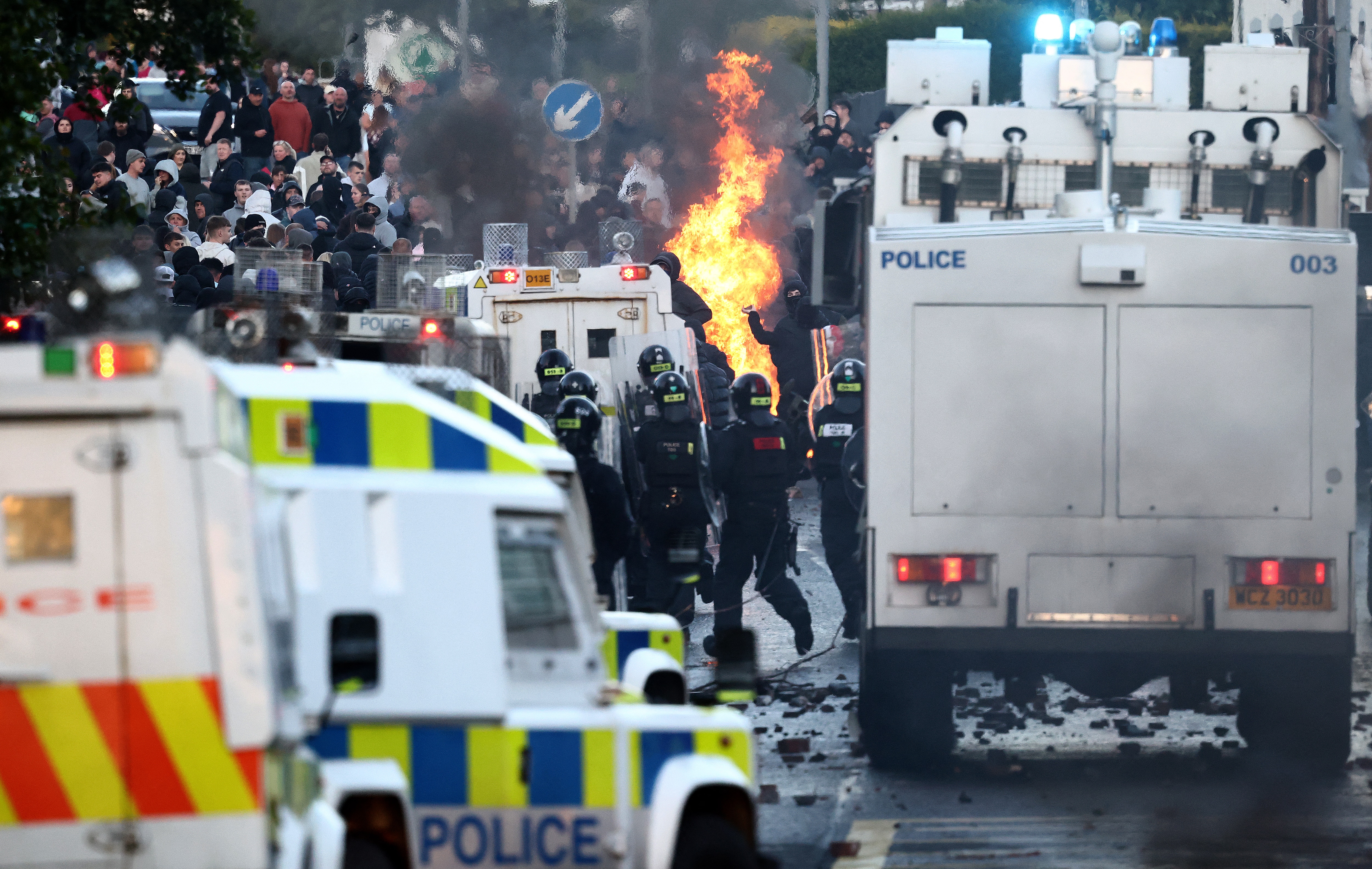 Police in black riot gear with white police vans confront a mass of people throwing projectiles in front of a fire.