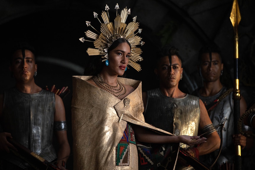 A woman wears an Incan gold head piece and cape and stands between men wearing armour, one holds a spear.