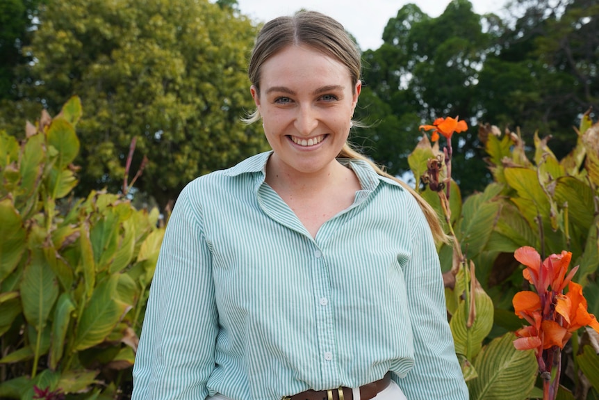 A woman in a green button up shirt standing in front of a garden smiles at the camera