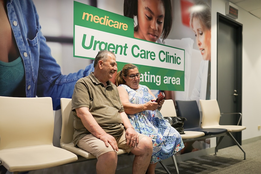 An older couple sit side by side in a waiting room, in front of a large sign that says "Medicare Urgent Care Clinic".