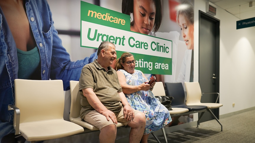 An older couple sit side by side in a waiting room, in front of a large sign that says "Medicare Urgent Care Clinic".