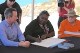 An Indigenous woman is filmed signing an agreement alongside men in suits at a table in an outdoor setting.